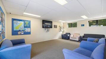 Une salle d’attente avec des canapés bleus, un téléviseur à écran plat, un tableau d’affichage et une fenêtre donnant sur un espace verdoyant.