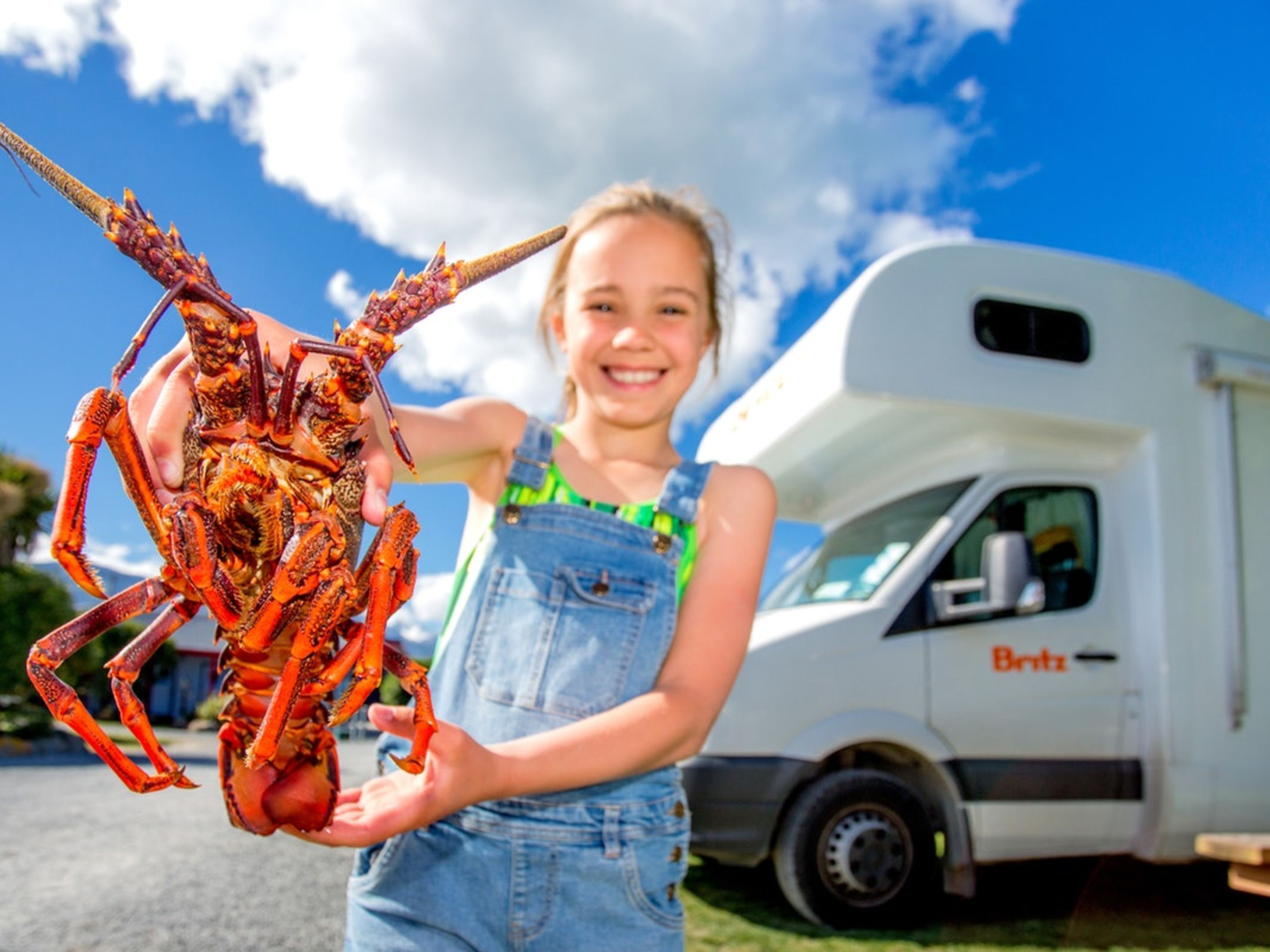 Una persona sostiene un langostino gigante frente a un vehículo blanco con el nombre de la marca 