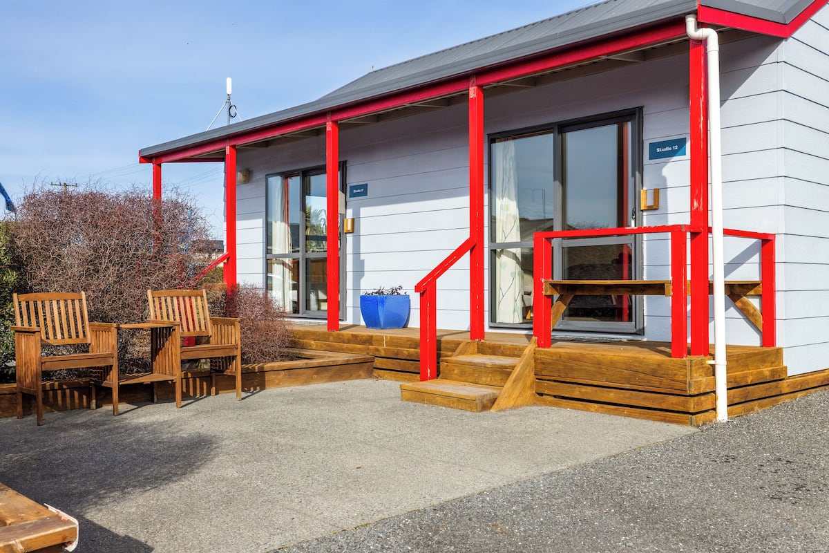 Un bâtiment à l’extérieur rouge et blanc, avec des marches en bois menant à une porte et un jardinier bleu.