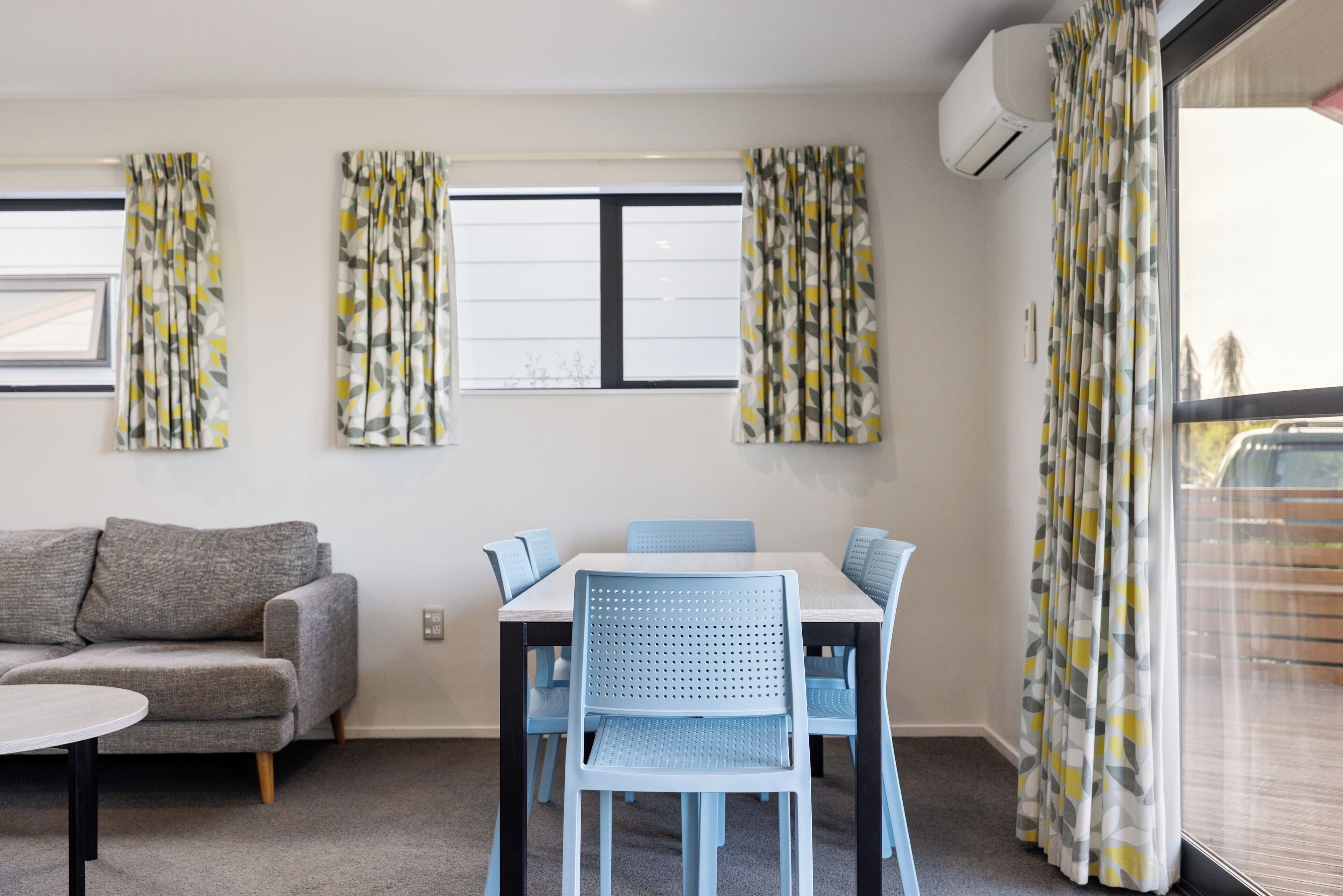 Una sala de estar moderna con un sofá gris, una mesa blanca y sillas azules. Hay tres ventanas con cortinas estampadas y un equipo de aire acondicionado en la pared.