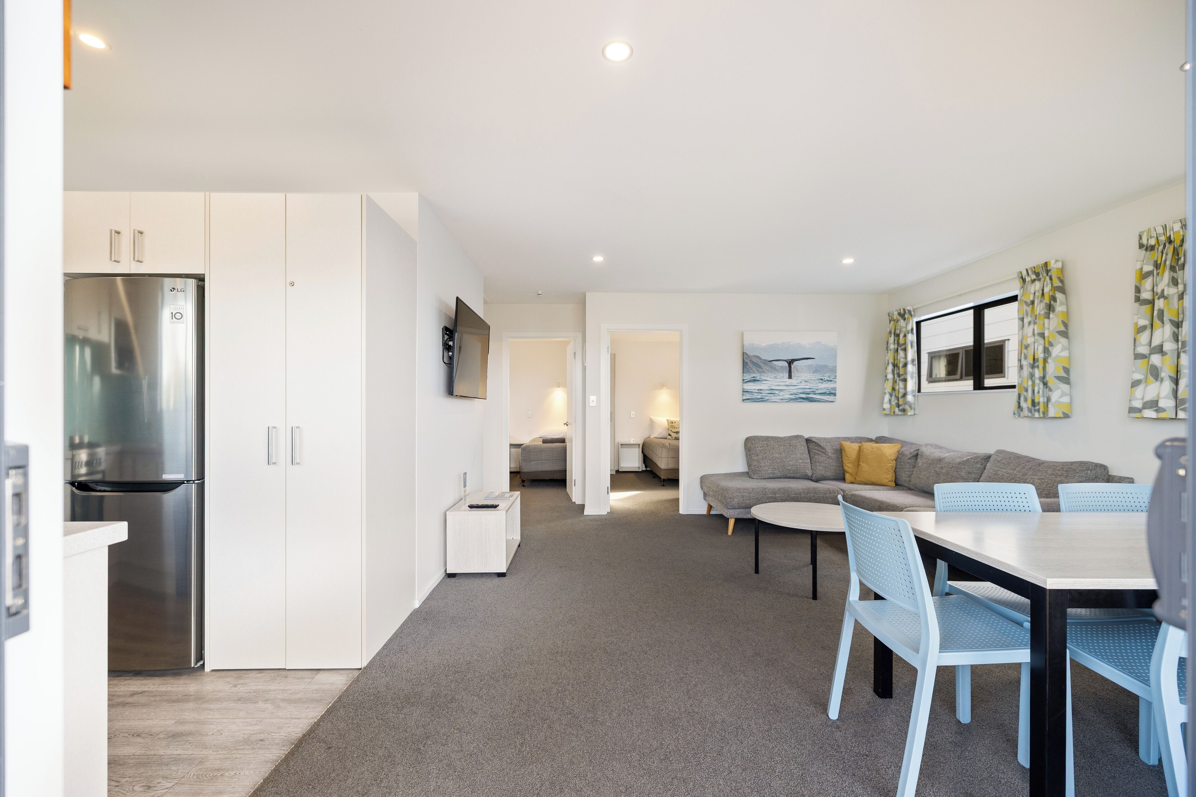 Een moderne keuken met witte kastjes en een roestvrijstalen koelkast. Er is een zithoek met een tafel en stoelen, en een woonkamer met een bank en een schilderij van een walvis.