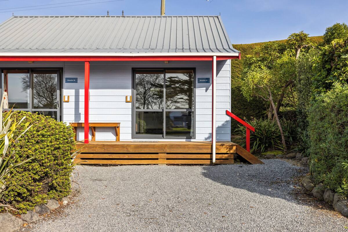 Un petit bâtiment d’un seul étage, à la façade rouge et blanche, avec un banc en bois à l’extérieur et une allée gravillonnée.