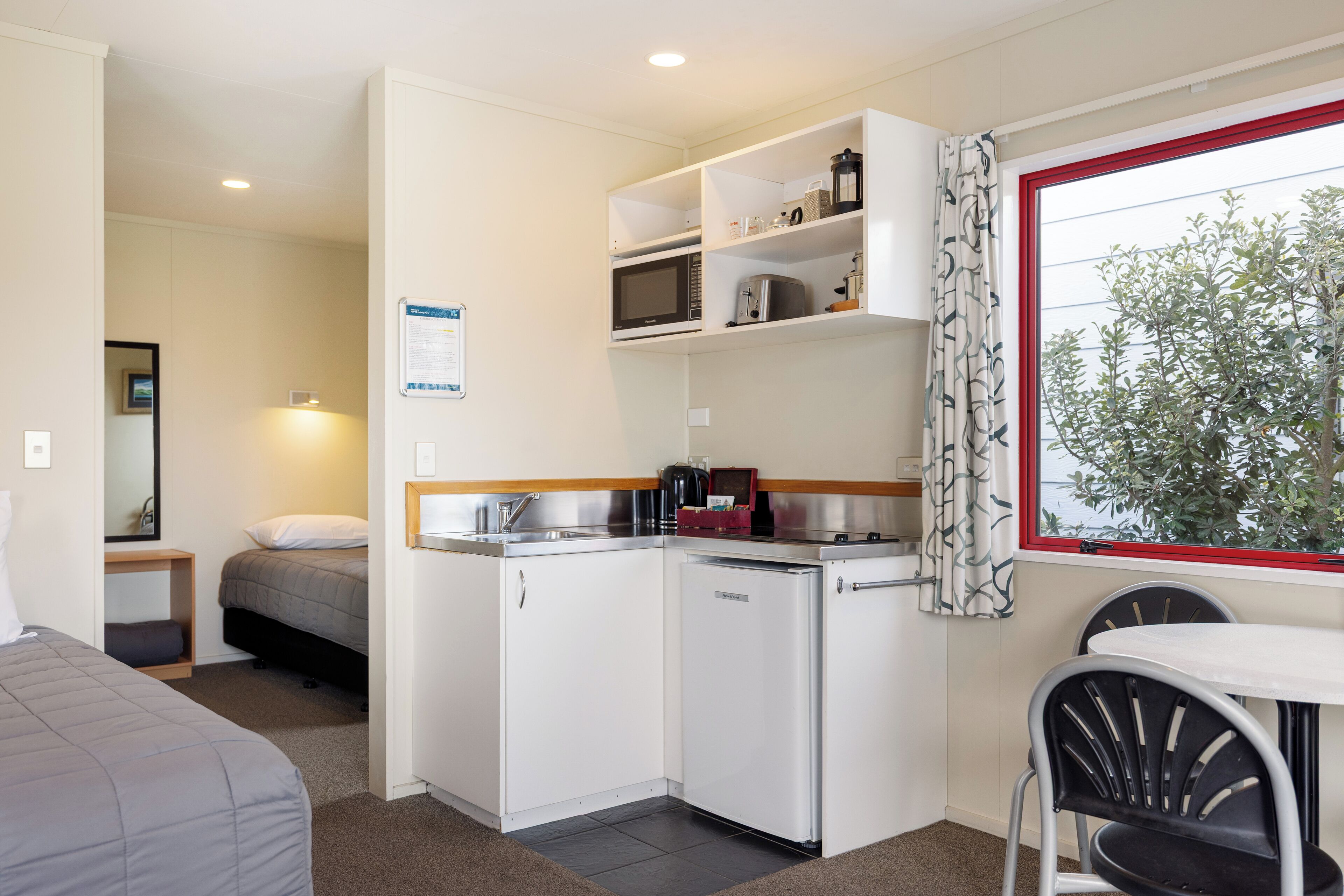 Een kleine hotelkamer met kitchenette, een bed, een tafeltje en een raam met uitzicht op bomen.