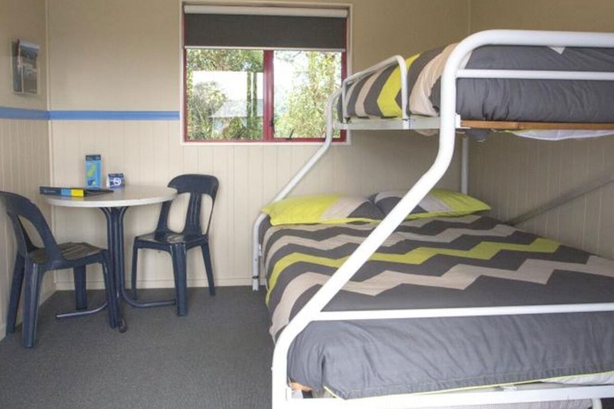 Een stapelbed met een bureau en twee stoelen, een raam met uitzicht op groen, en een wandplank met spullen erop.