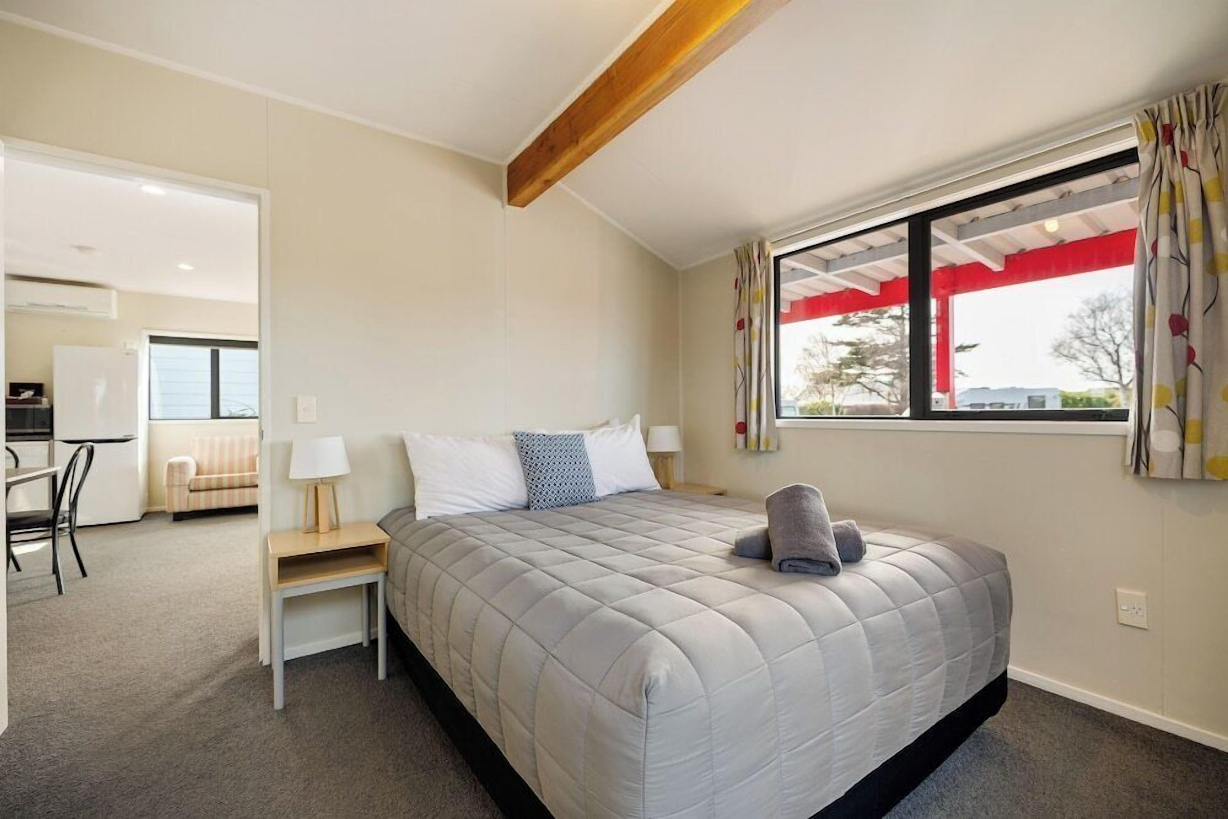 Une chambre à coucher avec un lit, une table de chevet, une lampe, une fenêtre avec des rideaux et une vue sur l’extérieur.