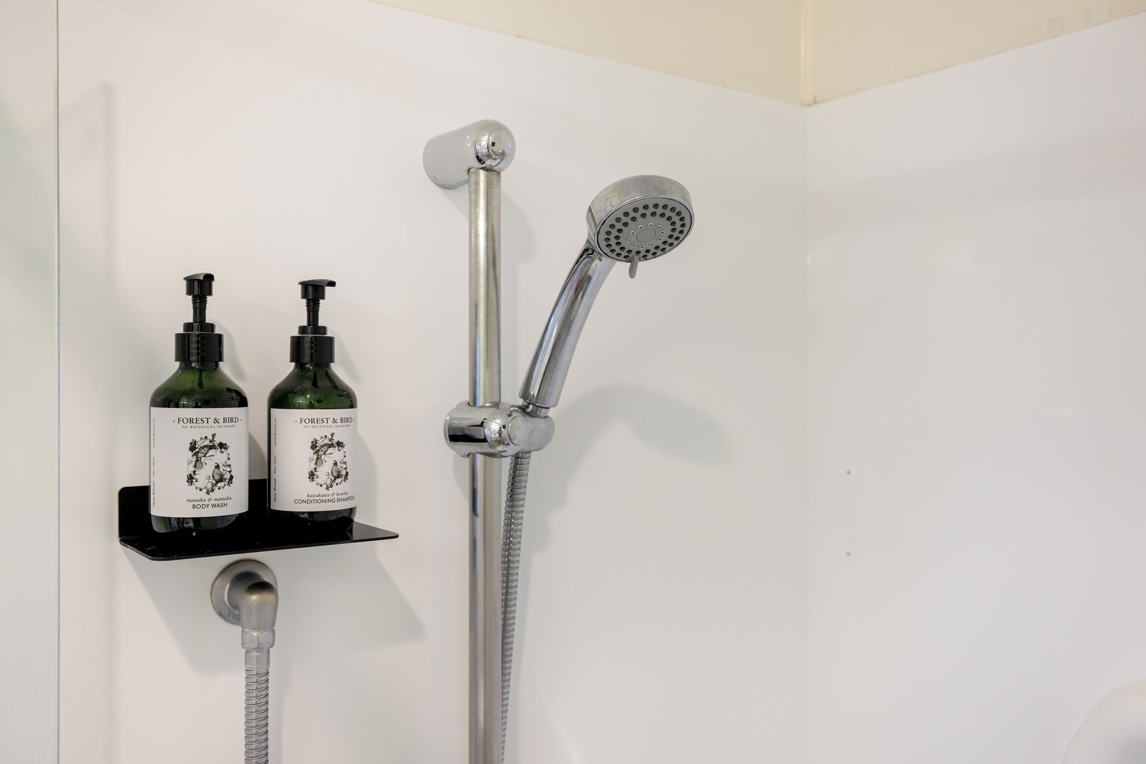 Ducha con cabezal de ducha de mano y grifo empotrado. Dos botellas de gel de baño y champú acondicionador están colocadas en un estante negro debajo de la ducha.