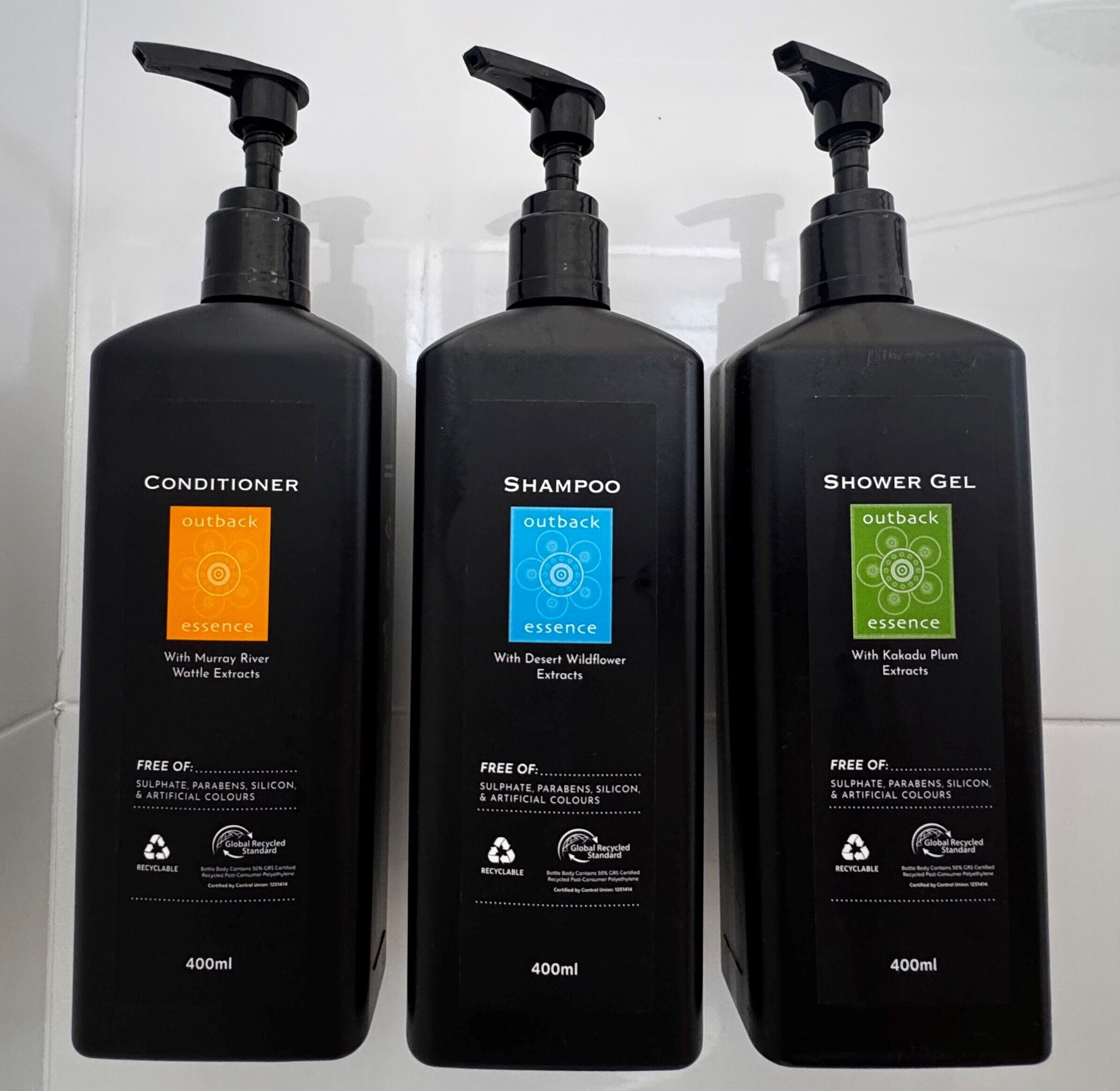 Tres botellas negras de productos de cuidado personal Outback Essence: acondicionador, champú y gel de ducha. Cada botella tiene 400ml y lleva la marca indicando que es reciclable.