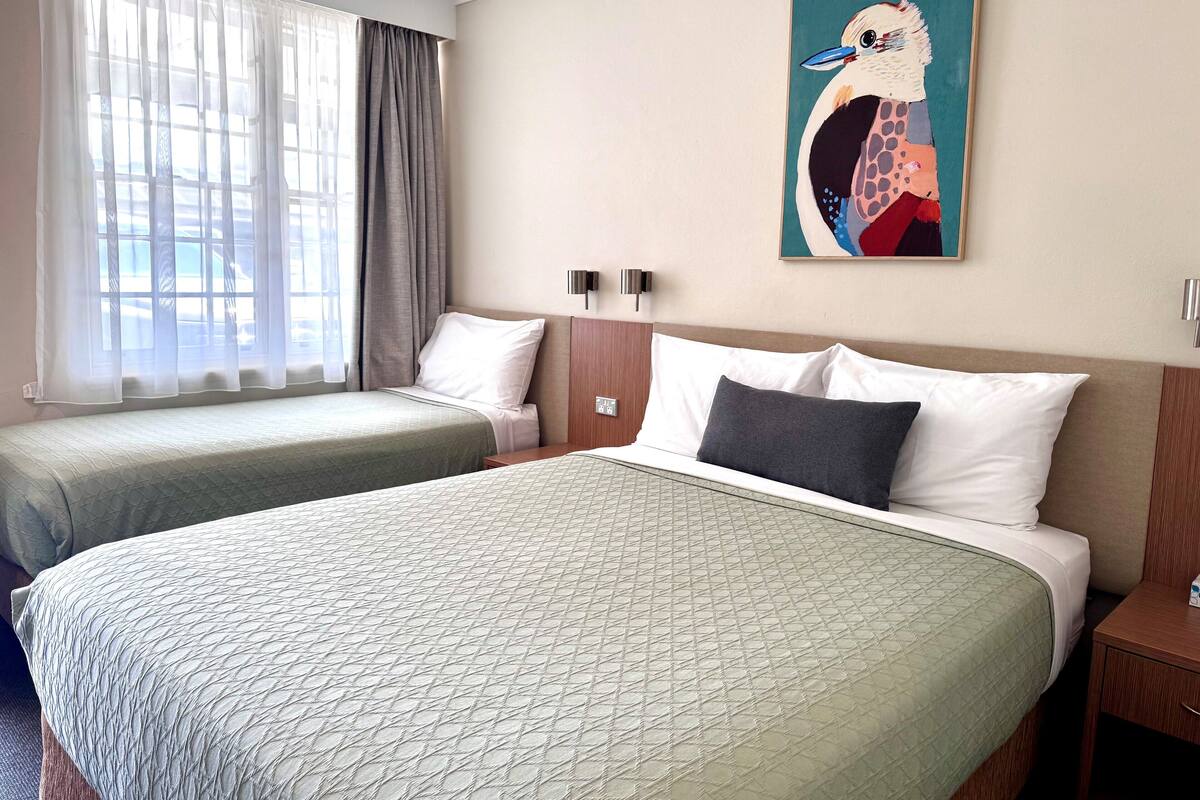 A hotel room with two beds, a large window with sheer curtains, a wall-mounted painting, and a bedside table.