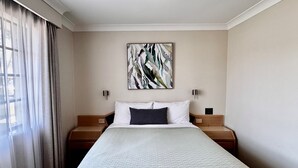 Habitación de hotel con una cama grande, dos mesitas de noche, un cuadro abstracto colgado en la pared y cortinas traslúcidas en la ventana.