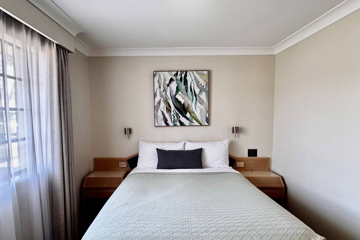 A hotel room with a large bed, two bedside tables, a wall-mounted abstract painting, and sheer curtains on the window.