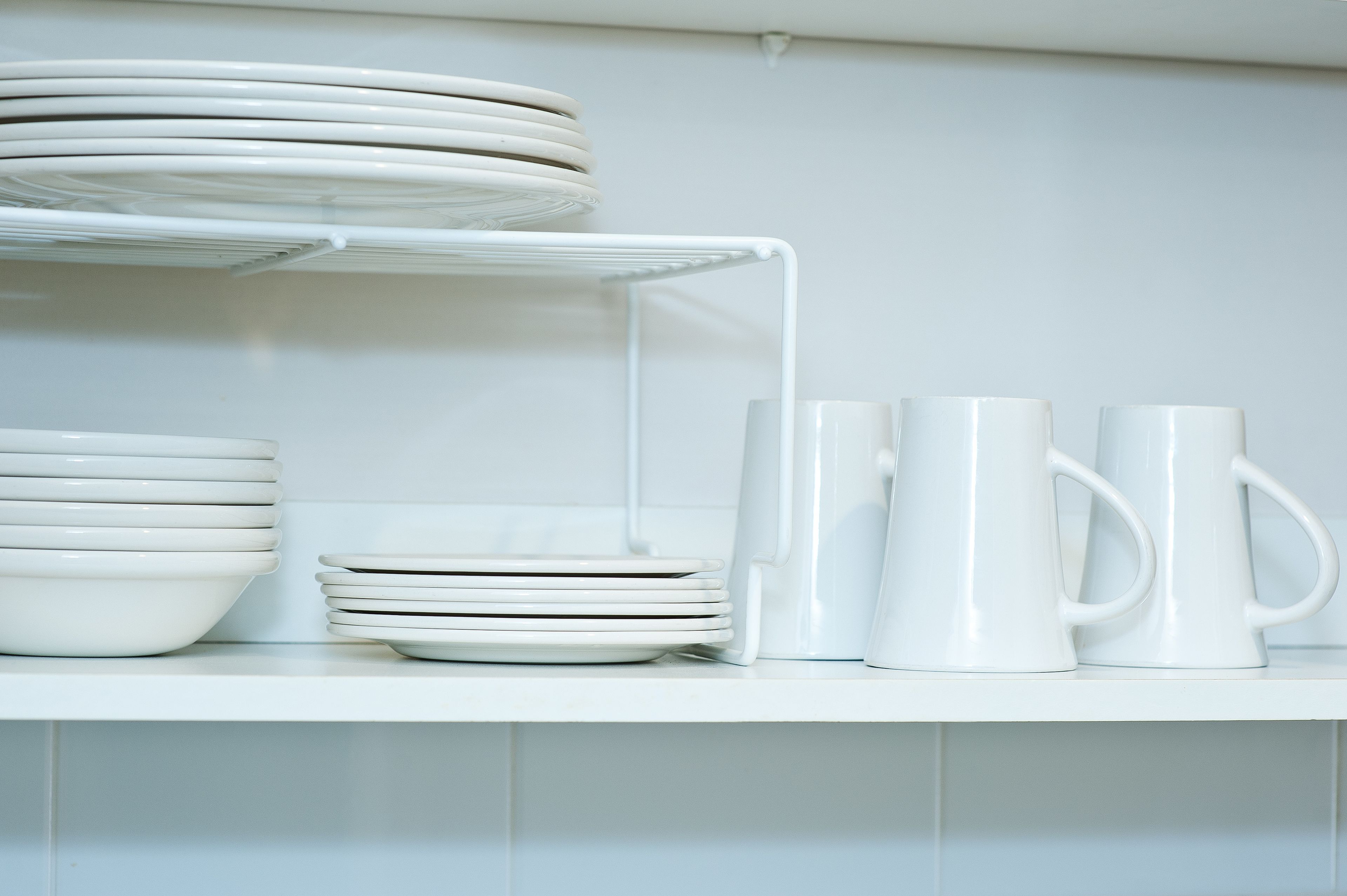A white shelf with stacked plates, bowls, and white mugs.