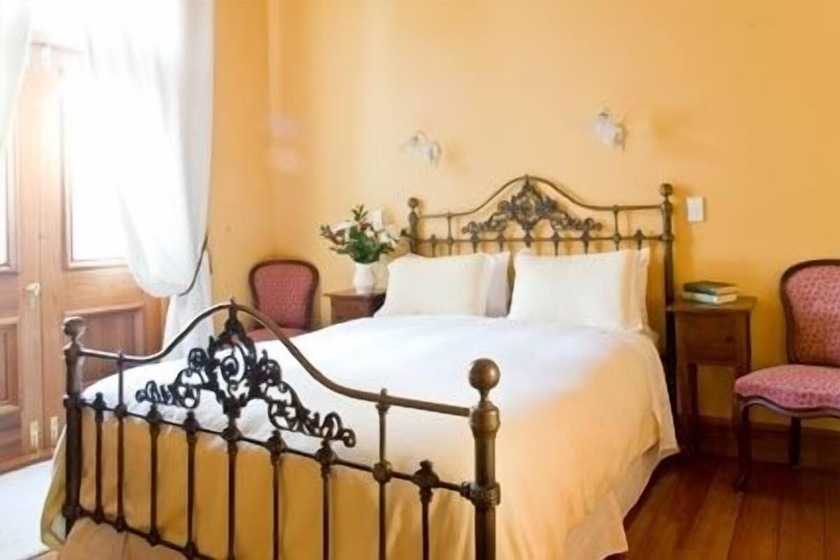 A neatly made bed with a metal headboard, a bedside table with a lamp, and a chair with a cushion.