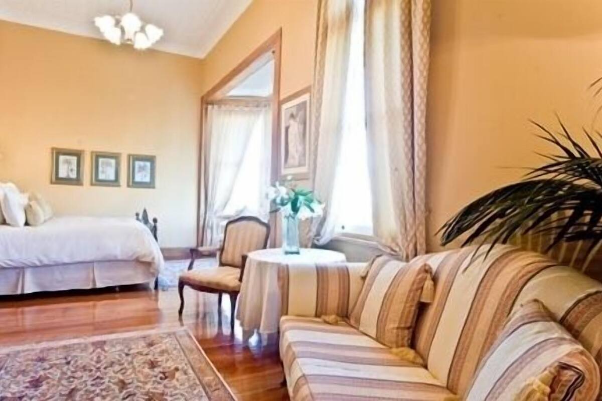 A hotel room with a bed, a striped sofa, a small round table with a vase, and a large window with curtains.