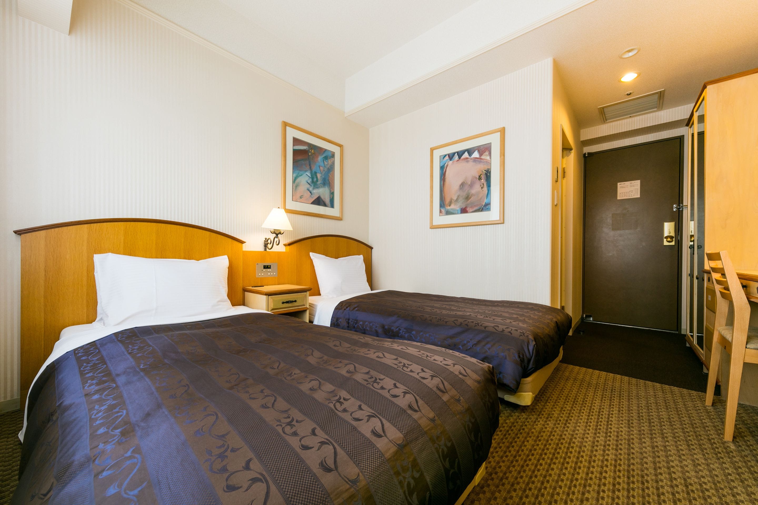 Een hotelkamer met twee bedden, een nachtkastje, een lamp en twee ingelijste foto’s aan de muur.
