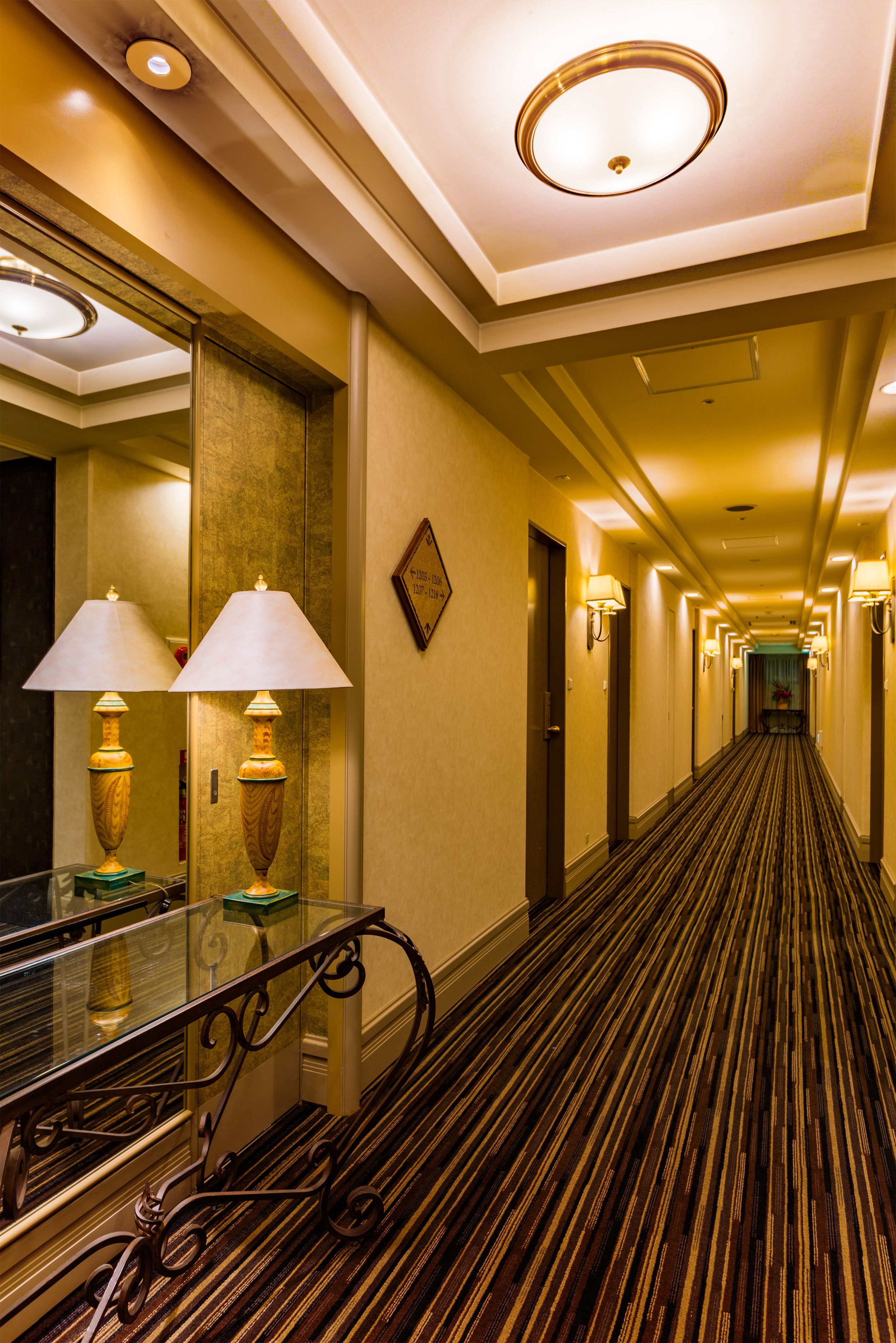 A hotel hallway with a carpeted floor, wall-mounted lamps, and a glass railing.