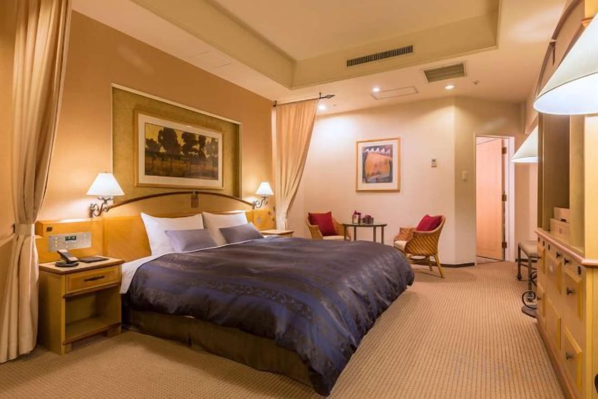 A hotel room with a bed, bedside lamps, a desk, a chair, a television, and a painting on the wall.