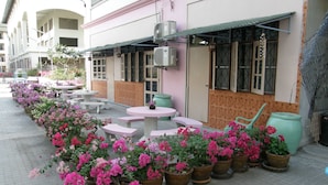 A row of outdoor tables and chairs along a building's exterior, with potted plants and flowers in the foreground. - Anchana House by Zuzu (Pattaya)