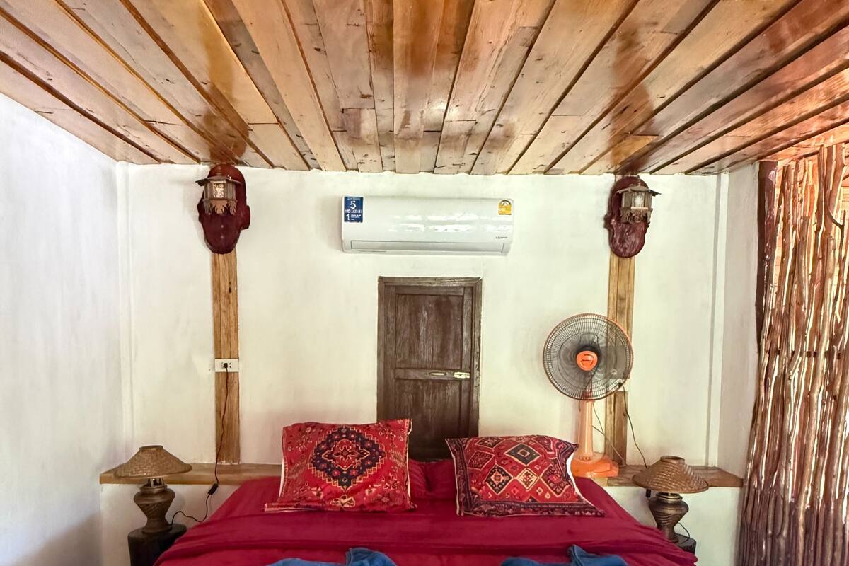 A room with a wooden ceiling, a bed with red bedding, a wall-mounted air conditioner, a door, and a fan.