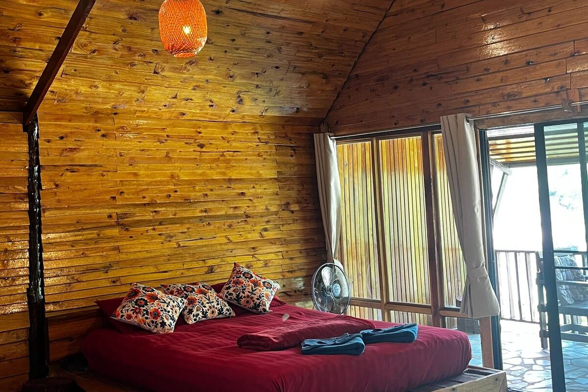 A wooden cabin interior with a bed, hanging orange lanterns, and a large window.