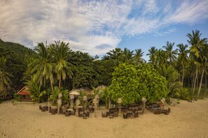 Un resort frente a la playa con asientos al aire libre, palmeras y una edificación con techo de paja.