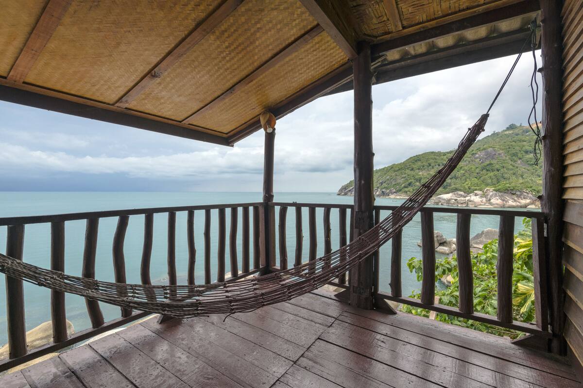 A wooden deck with a hammock, overlooking a body of water and a mountainous landscape.