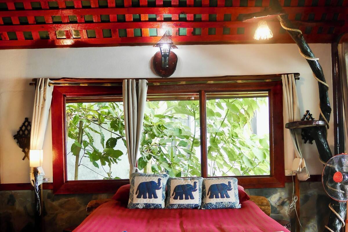 A room with a large bed, a window with curtains, a hanging light fixture, and a view of greenery outside.