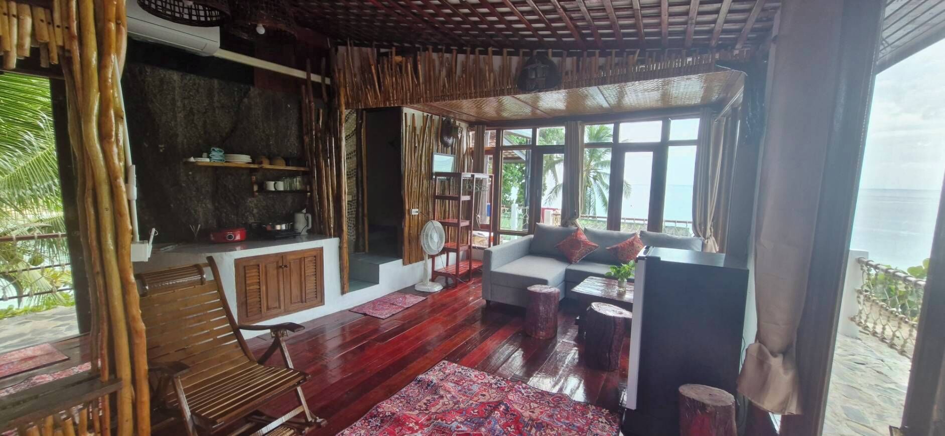A room with a wooden floor, a sofa, a television, and a window with a view of the sea.