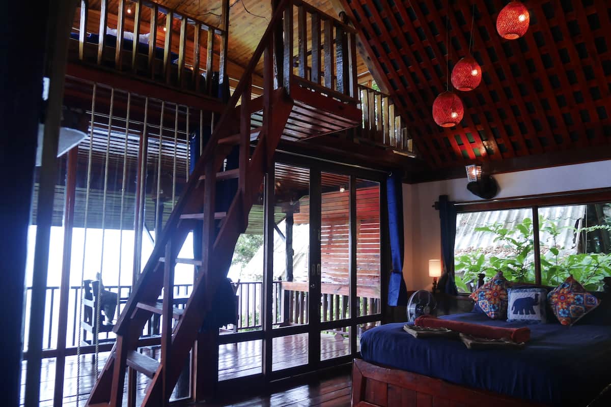 A room with a wooden staircase, a bed with colorful pillows, and a view of greenery outside.