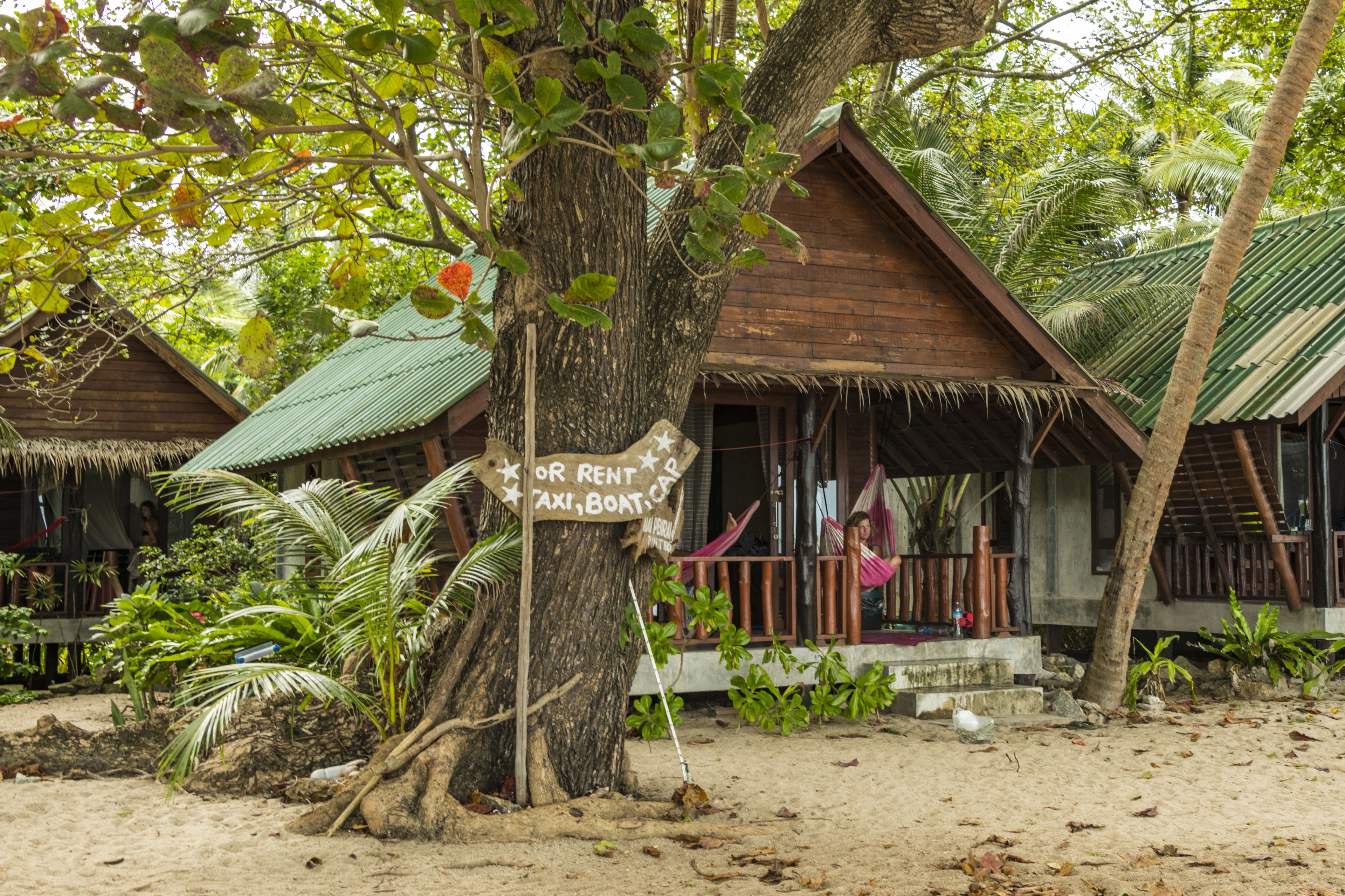 A wooden bungalow with a thatched roof, a porch, and a sign indicating it's for rent.