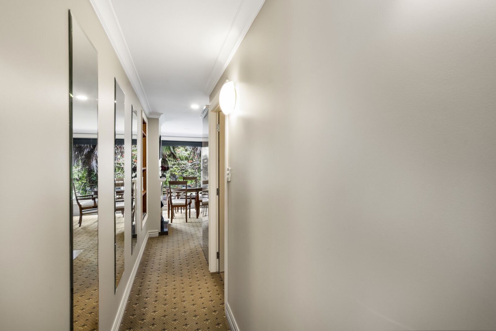 A hallway with a patterned carpet, white walls, and a view of an outdoor patio with a table and chairs.
