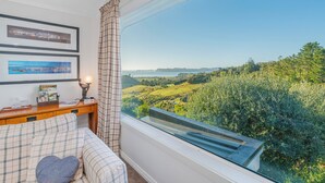 A bedroom with a large window overlooking a scenic landscape, a bed with plaid bedding, and a bedside table with a lamp and a book titled "COROMIND".