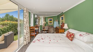 A bedroom with a large bed, a seating area with a sofa and chairs, a desk, and a view through a sliding door to a garden.
