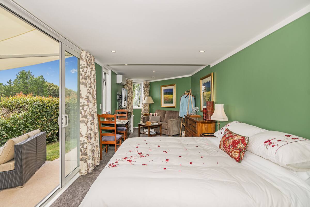 Ein Schlafzimmer mit einem großen Bett, einer Sitzecke mit Sofa und Sesseln, einem Schreibtisch und Blick durch eine Schiebetür auf einen Garten.