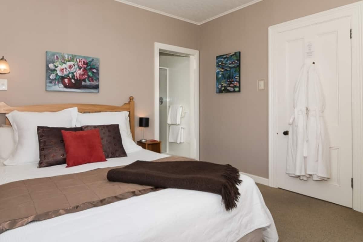 Quarto com cama de madeira, roupa de cama branca e castanha, duas pinturas, um roupão branco pendurado numa porta e uma mesa de cabeceira com uma lâmpada.