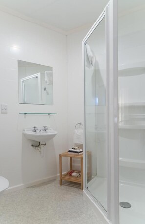 Un bany modern amb una cabina de dutxa de vidre, un lavabo blanc amb un mirall a sobre i un petit tamboret de fusta.