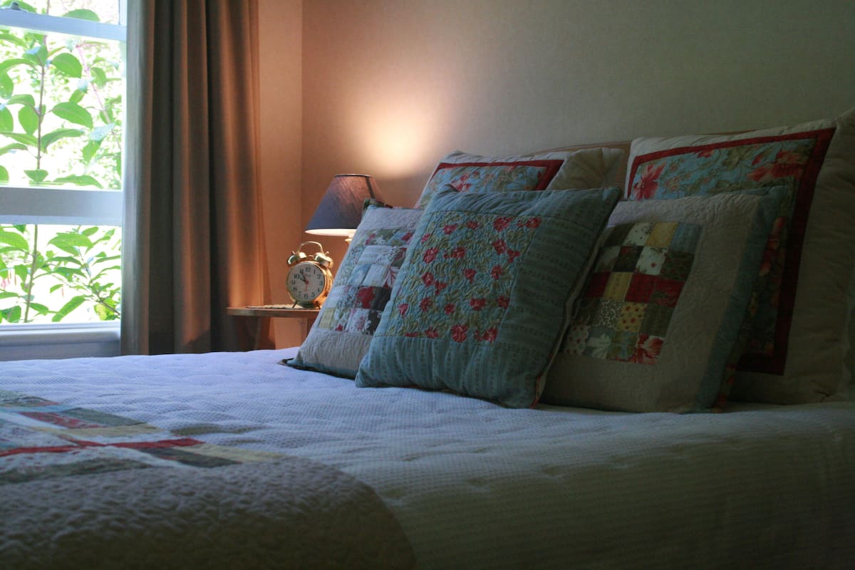 Cama bem arrumada com colcha e várias almofadas, mesa de cabeceira com abajur e janela com cortinas.