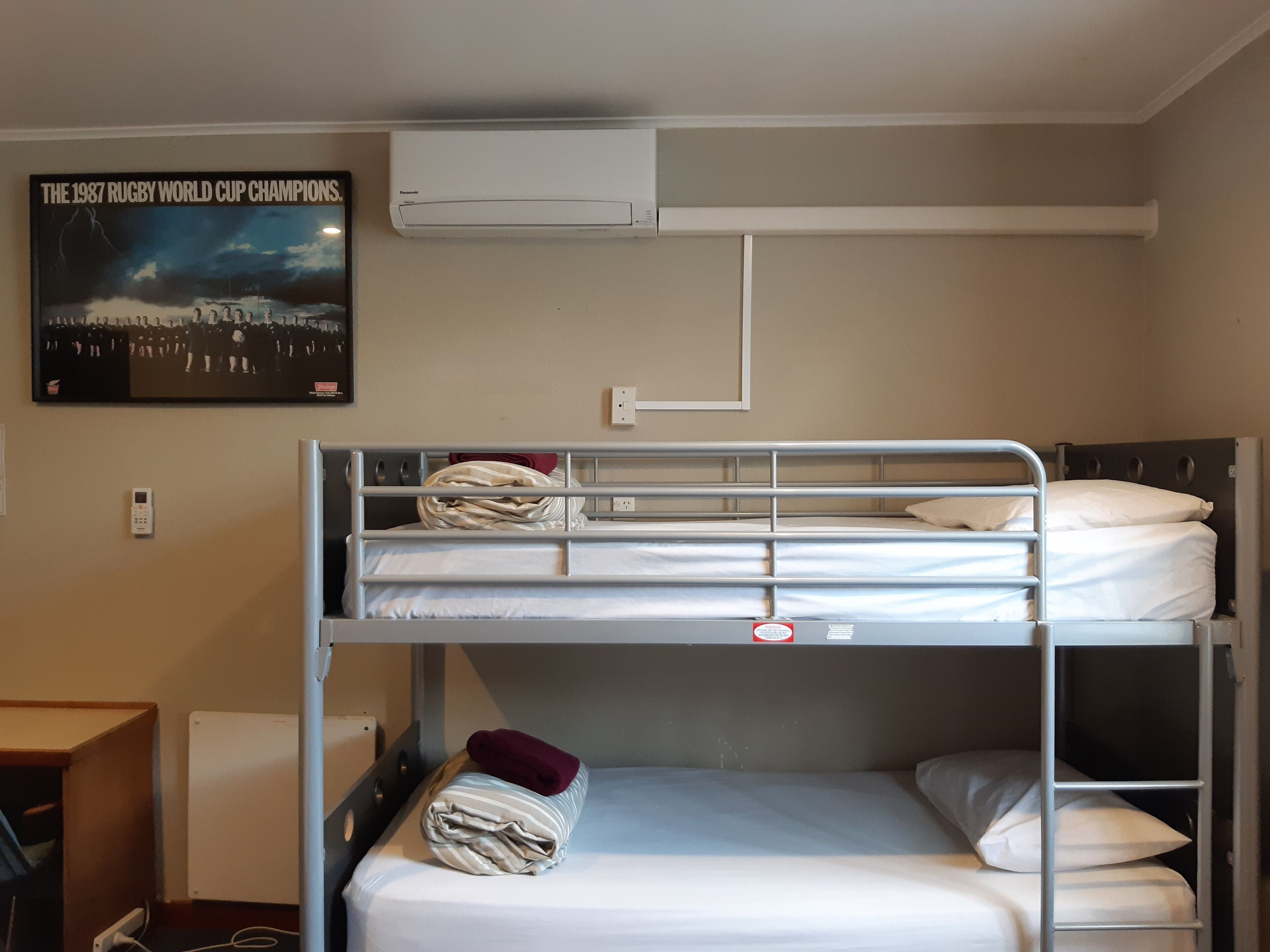 Habitación con literas, un cartel del equipo campeón de la Copa Mundial de Rugby de 1987, un aire acondicionado y un escritorio con silla.
