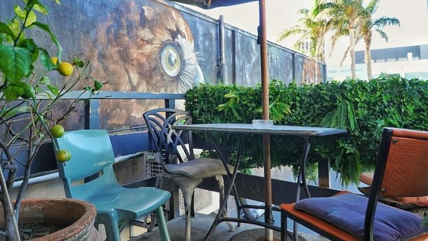 Uma área externa com mesas e cadeiras, um mural na parede e vista para palmeiras.
