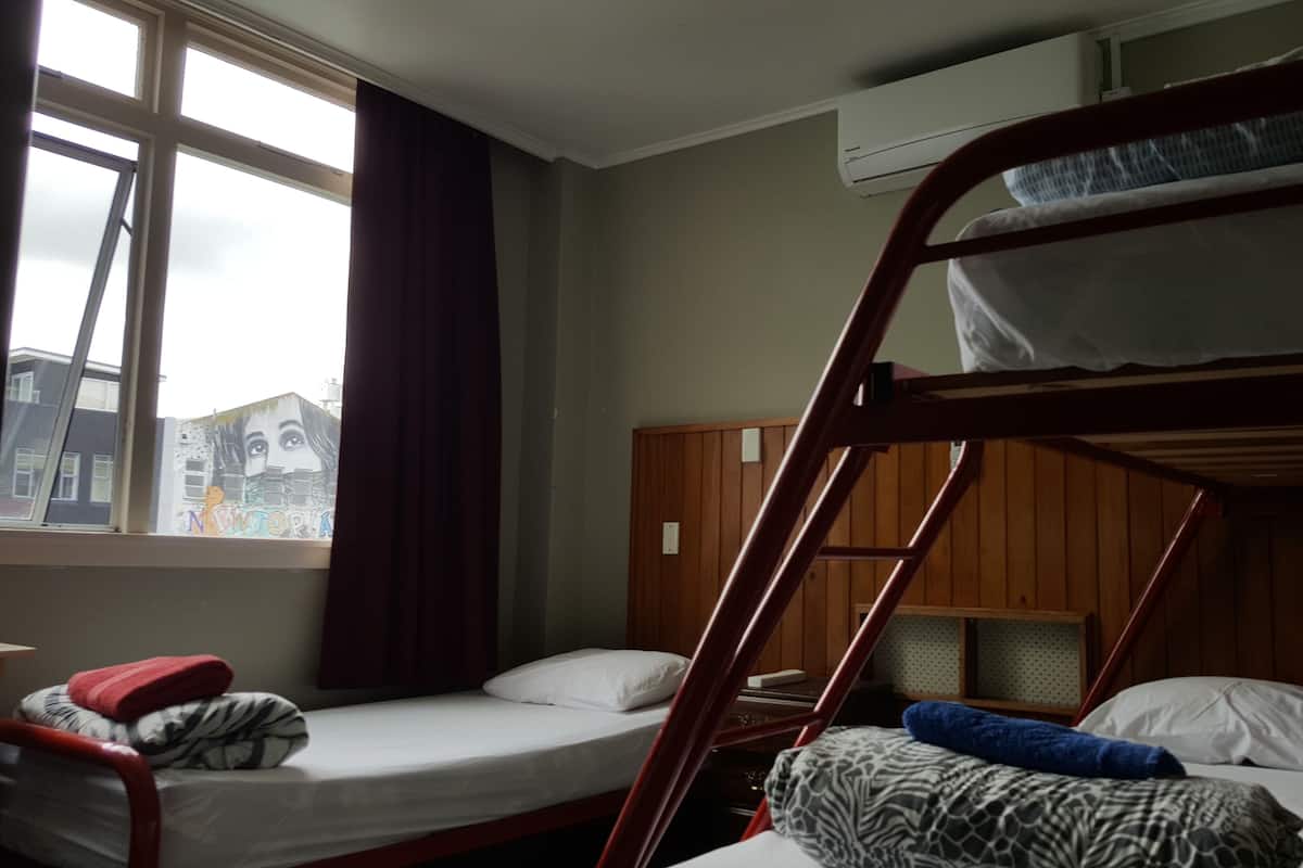 A dormitory room with bunk beds, a window with curtains, and a view of buildings outside.