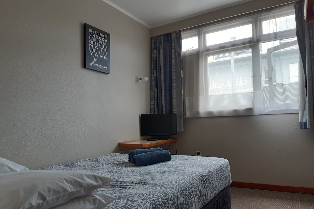 A bedroom with a bed, a television on a wooden nightstand, a window with curtains, and a wall with a framed sign.