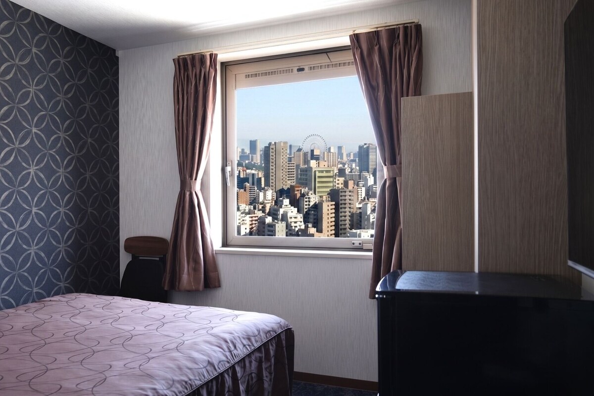 A hotel room with a bed, a small table, a window with curtains, and a cityscape view.