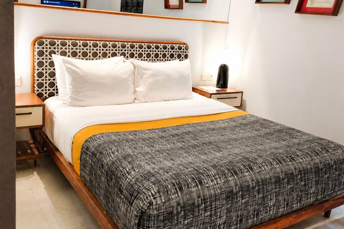 A bedroom with a wooden bed frame, white bedding, a patterned throw blanket, a bedside table with a lamp, and a wall-mounted air conditioner.
