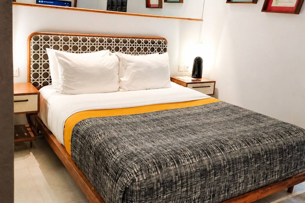 A bedroom with a wooden bed frame, white bedding, a patterned throw blanket, a bedside table with a lamp, and a wall-mounted air conditioner.