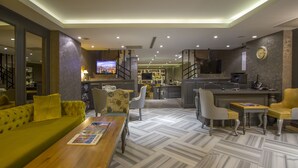 A modern hotel lobby with a sofa, chairs, a wooden table, a kitchen area, and a TV displaying a cityscape. - Aston Hotel Taksim (Istanbul)