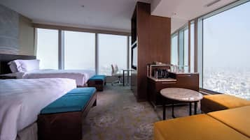 A hotel room with two beds, a desk, a chair, a small table, and a large window offering a city view.