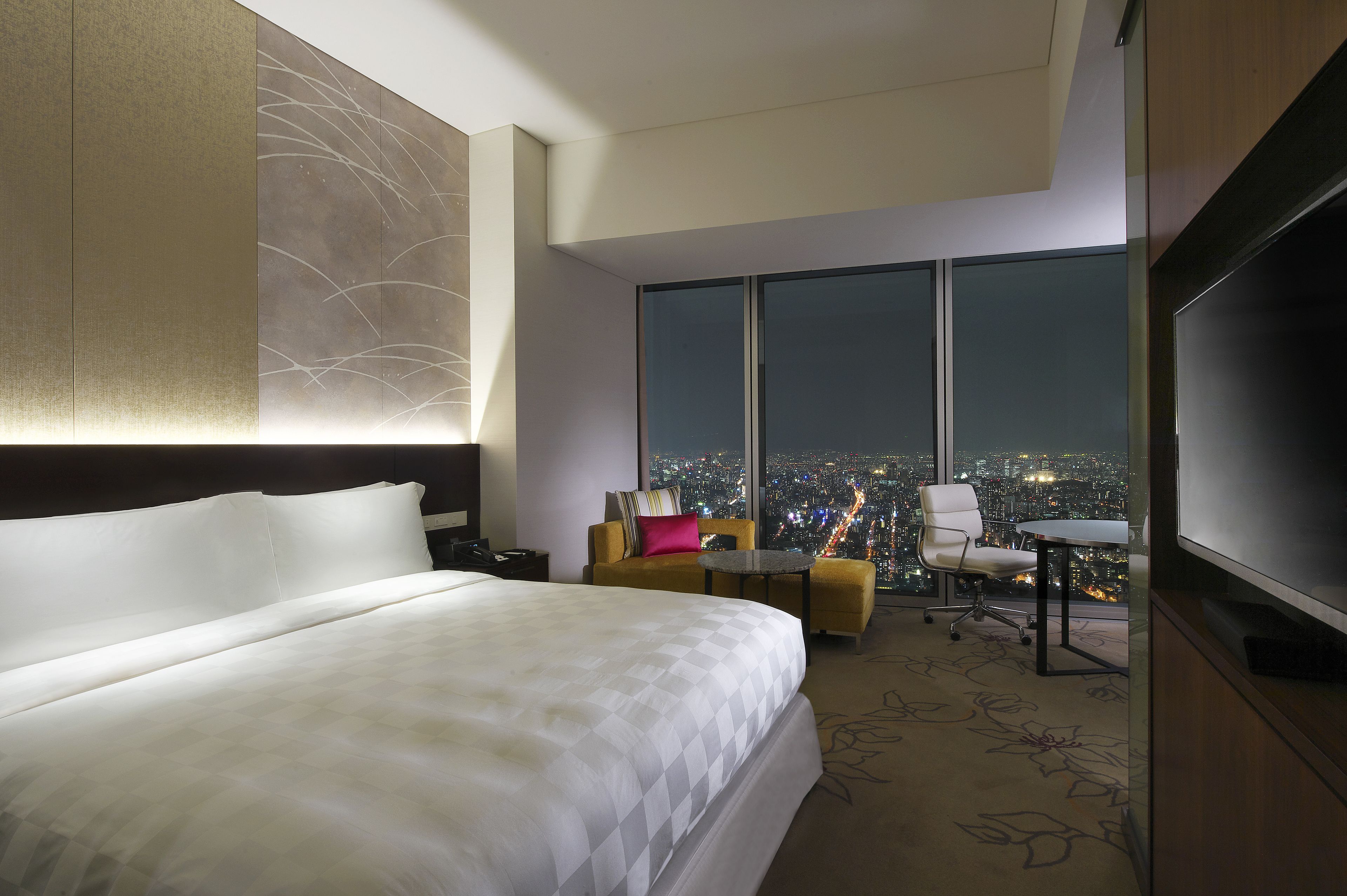 A hotel room with a large bed, a desk with a chair, a television, and a view of a cityscape at night.