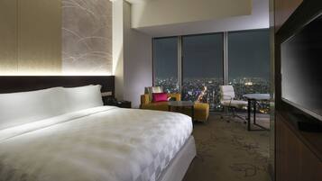 A hotel room with a large bed, a desk with a chair, a television, and a view of a cityscape at night.