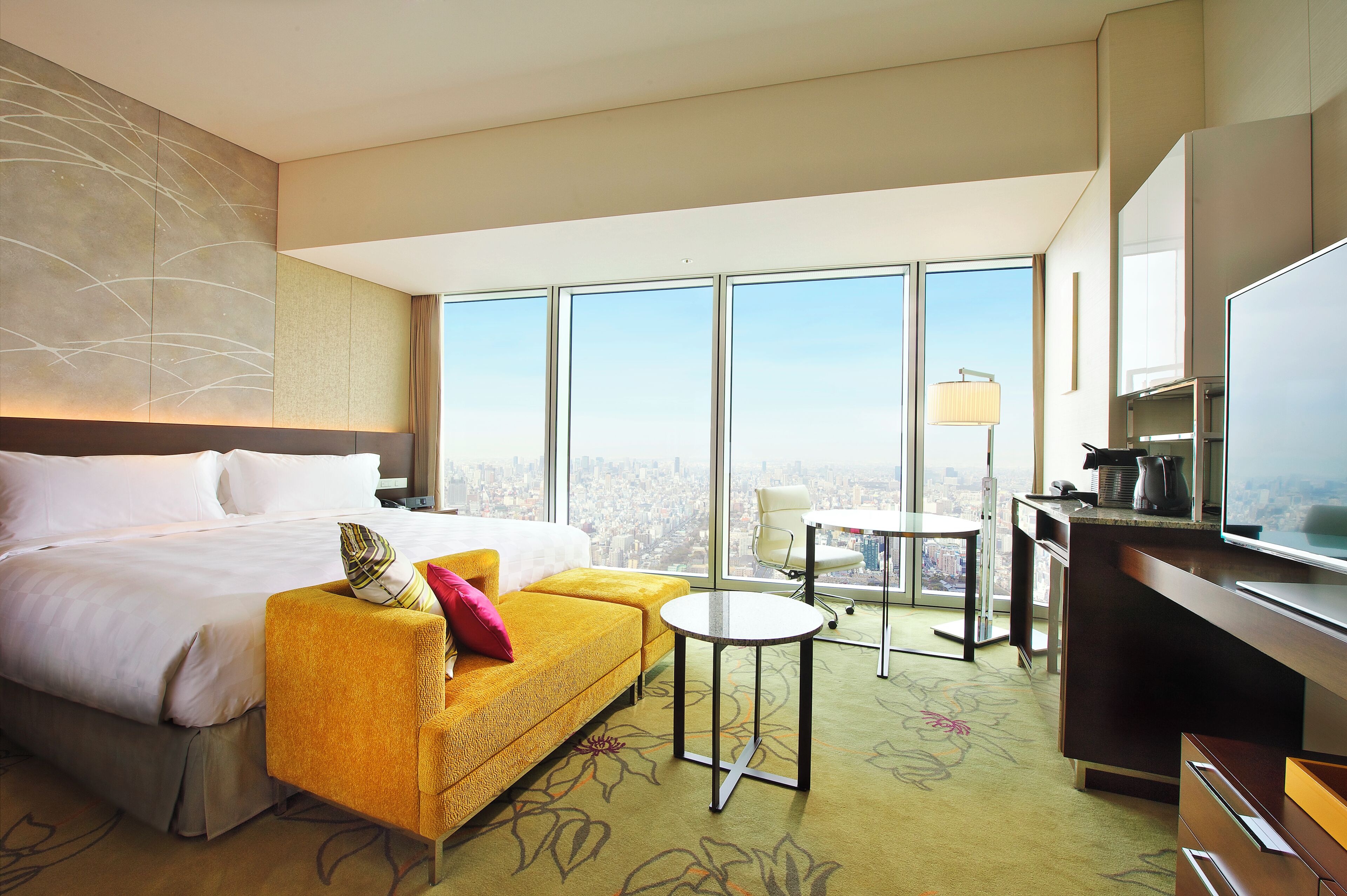 A hotel room with a large bed, a yellow sofa, a desk with a TV, and a city view through large windows.