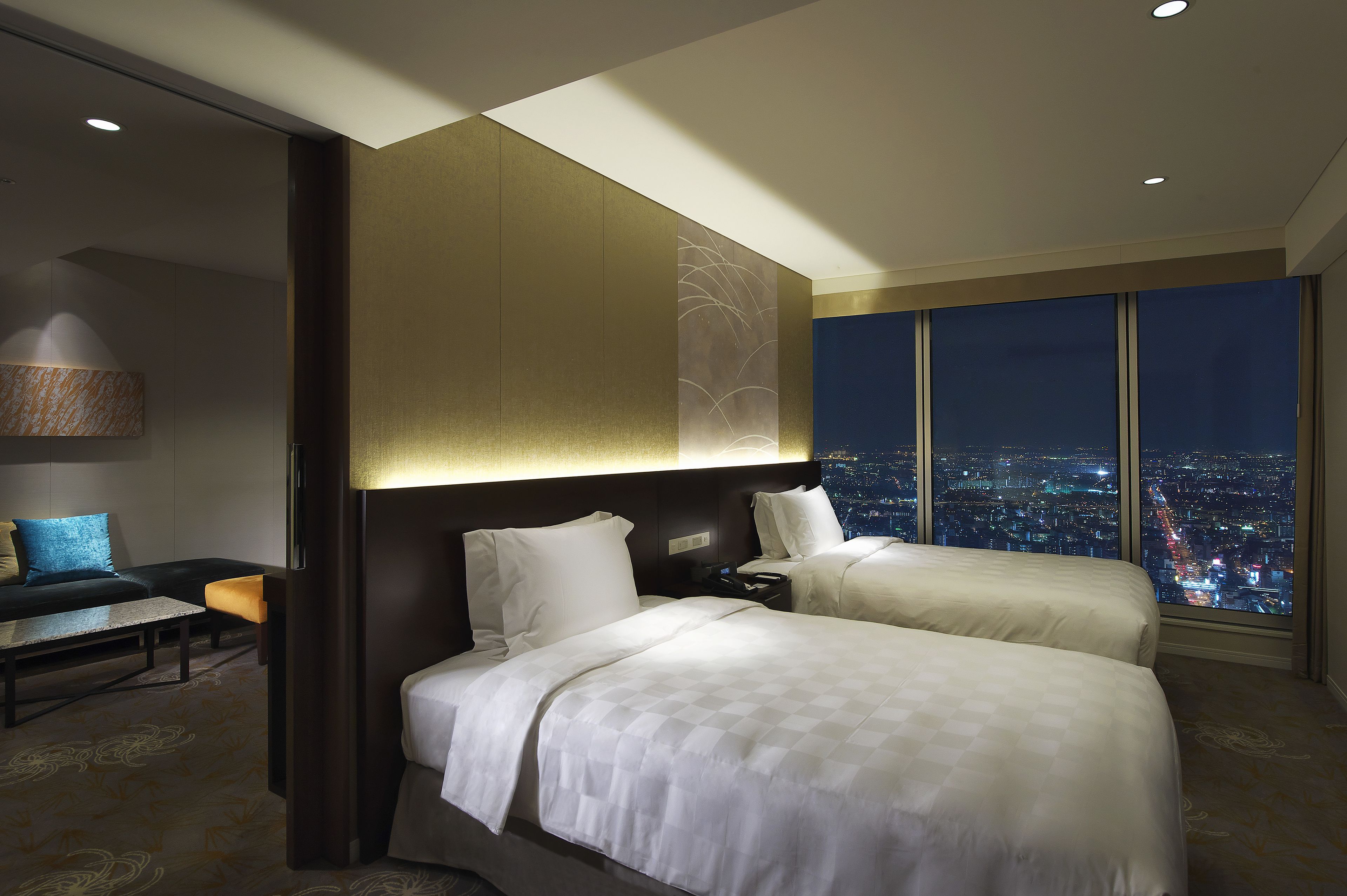 A hotel room with two beds, a desk, a chair, and a large window with a city view.