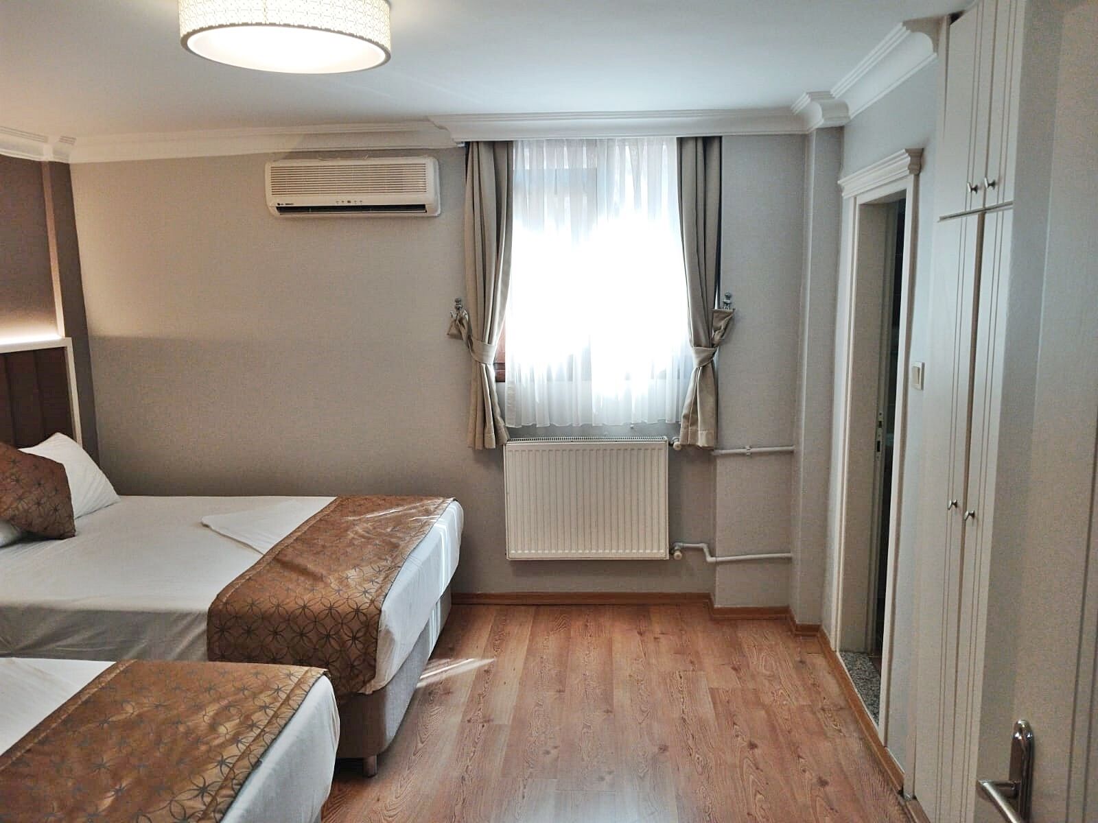 Quarto de hotel com duas camas, um radiador, uma janela com cortinas e um guarda-roupa.